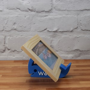 May include: A blue whale-shaped picture frame stand holds a wooden picture frame with a photo of a beach scene. The stand has the word "We" printed on it.