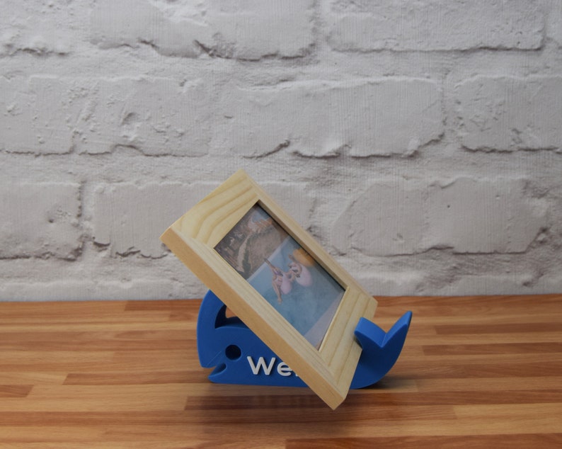 May include: A blue whale-shaped picture frame stand holds a wooden picture frame with a photo of a beach scene. The stand has the word "We" printed on it.