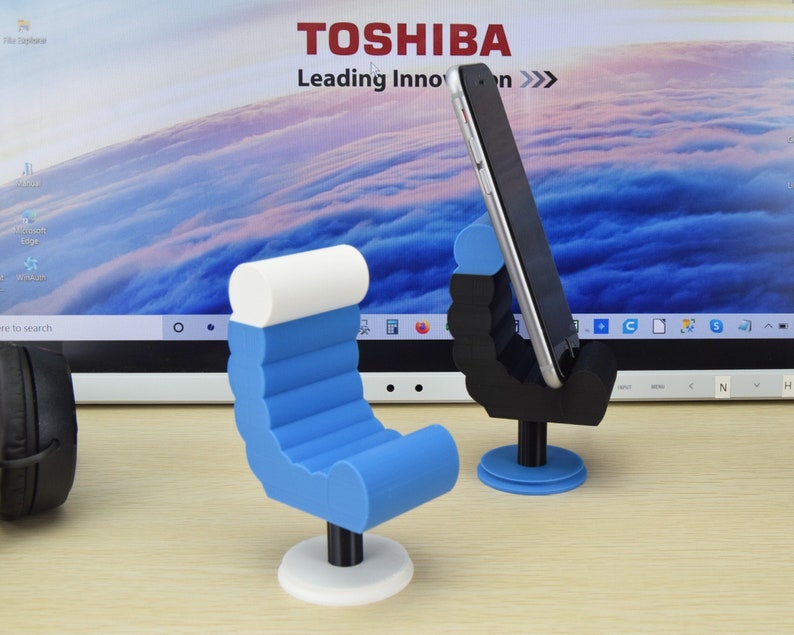 May include: Two phone stands, one shaped like a chair and the other shaped like a hand, both in blue and white. The chair stand has a white base and the hand stand has a blue base. Both stands are on a wooden surface.