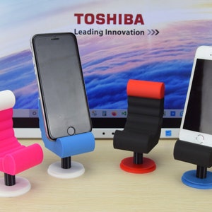 May include: Four phone stands in the shape of chairs. The stands are made of black, red, blue, and pink plastic. The stands are on a wooden surface. The stands are designed to hold a smartphone upright.