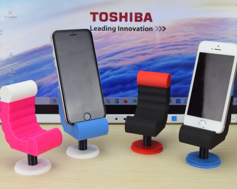 May include: Four phone stands in the shape of chairs. The stands are made of black, red, blue, and pink plastic. The stands are on a wooden surface. The stands are designed to hold a smartphone upright.