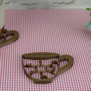 May include: A brown, cup-shaped coaster with the word "Caffeine" and a molecular structure design. The coaster sits on a red and white checkered surface, next to a matching mug and a small watering can.