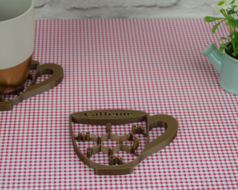 May include: A brown, cup-shaped coaster with the word "Caffeine" and a molecular structure design. The coaster sits on a red and white checkered surface, next to a matching mug and a small watering can.