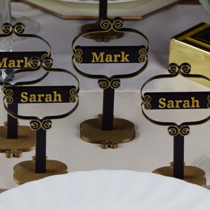 May include: Three black and gold table place card holders with the names Mark and Sarah written in gold lettering.