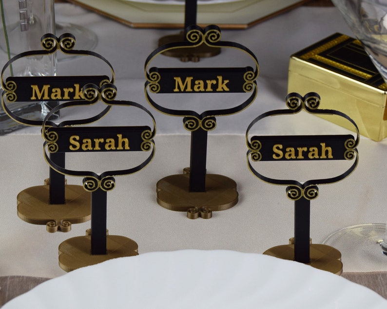 May include: Three black and gold table place card holders with the names Mark and Sarah written in gold lettering.