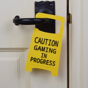 Op de afbeelding: Een gele deurhanger met de tekst "CAUTION GAMING IN PROGRESS".