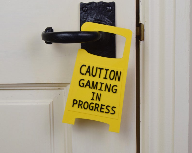 Op de afbeelding: Een gele deurhanger met de tekst "CAUTION GAMING IN PROGRESS".