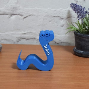 May include: A blue plastic snake with googly eyes and the name "John" printed on the side. The snake is curled up on a wooden surface.