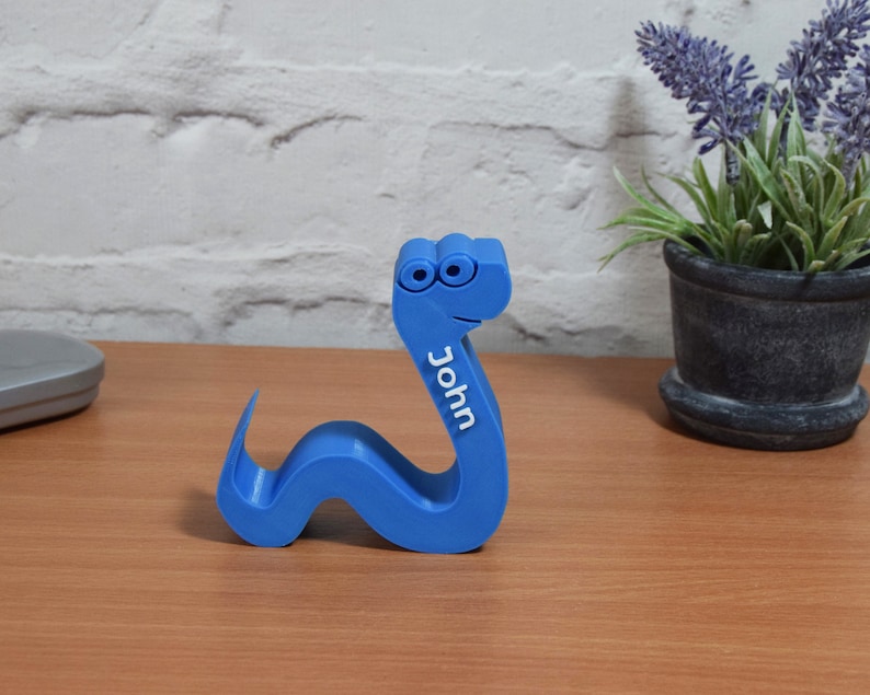 May include: A blue plastic snake with googly eyes and the name "John" printed on the side. The snake is curled up on a wooden surface.