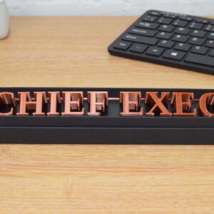 May include: A black box with a copper-colored sign that reads "CHIEF EXEC" in block letters.