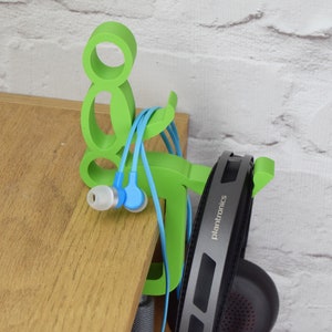 May include: Green plastic headphone and earbud holder shaped like a person. The holder has a hook for headphones and a slot for earbuds. The holder is sitting on a wooden surface.