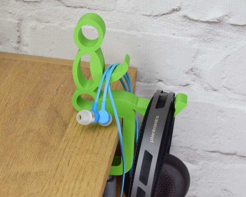 May include: Green plastic headphone and earbud holder shaped like a person. The holder has a hook for headphones and a slot for earbuds. The holder is sitting on a wooden surface.