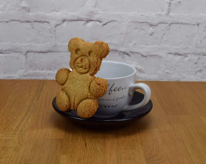 May include: A white ceramic coffee cup with the text "coffee... nice and frothy cappuccino" on the side. A brown bear-shaped cookie is resting on the rim of the cup.