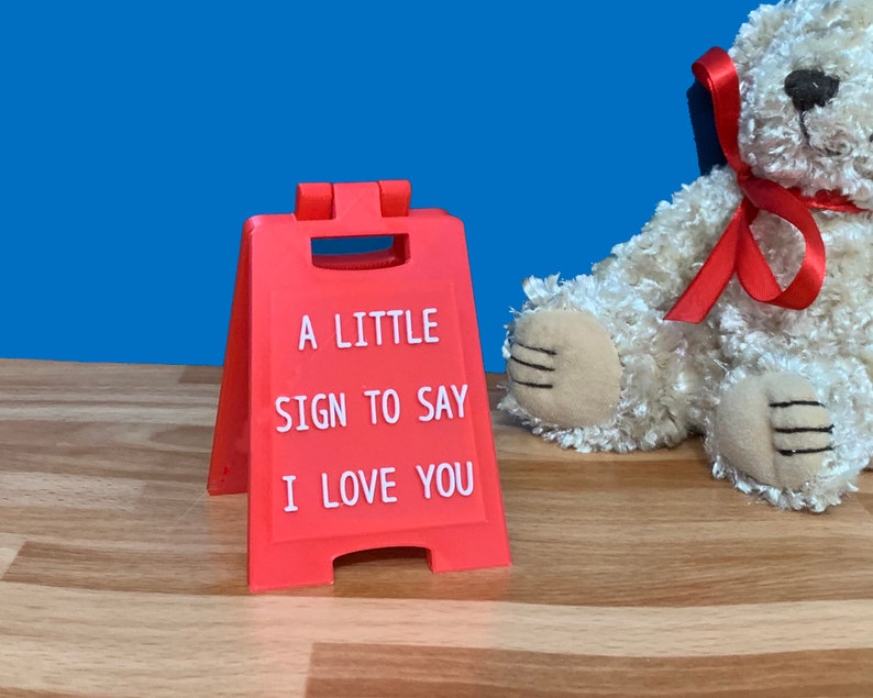 Op de afbeelding: Een rood plastic bord met de woorden "A little sign to say I love you" in witte letters. Het bord staat op een houten oppervlak naast een witte teddybeer met een rood lint.