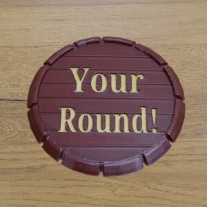 May include: A round, burgundy-colored sign with the words "Your Round!" in a lighter, gold-colored font. The sign is designed to resemble a barrel end, with horizontal lines and curved edges. The sign is on a wooden surface.