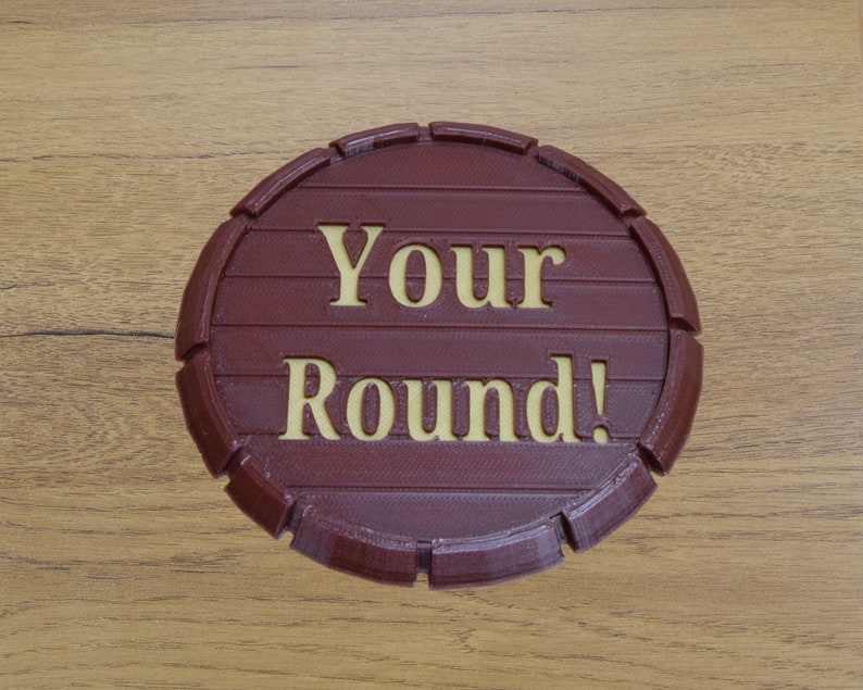 May include: A round, burgundy-colored sign with the words "Your Round!" in a lighter, gold-colored font. The sign is designed to resemble a barrel end, with horizontal lines and curved edges. The sign is on a wooden surface.