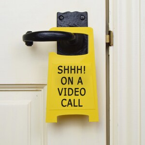 Op de afbeelding: Een geel bord met zwarte tekst dat "SHHH! ON A VIDEO CALL" zegt, hangt aan een witte deur.