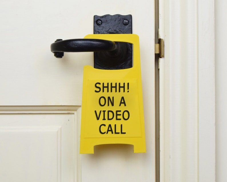 Op de afbeelding: Een geel bord met zwarte tekst dat "SHHH! ON A VIDEO CALL" zegt, hangt aan een witte deur.
