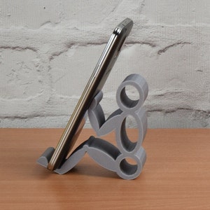 May include: A gray 3D printed phone stand shaped like a person holding a smartphone. The stand is on a wooden surface.