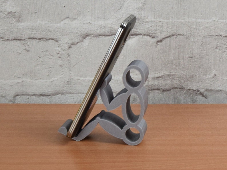 May include: A gray 3D printed phone stand shaped like a person holding a smartphone. The stand is on a wooden surface.