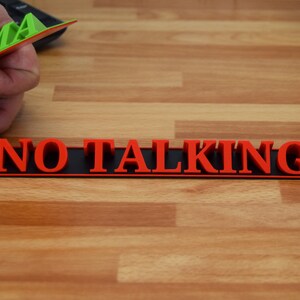 May include: A red "NO TALKING" sign with black backing is displayed on a wooden surface. A hand holds a green and red object with the letters "AMA" visible. The sign is designed to be a visual reminder.