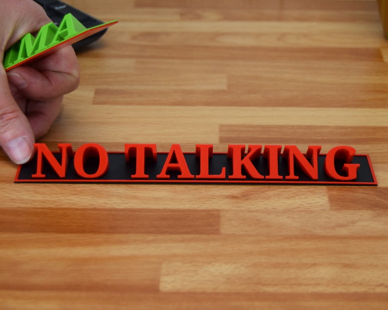 May include: A red "NO TALKING" sign with black backing is displayed on a wooden surface. A hand holds a green and red object with the letters "AMA" visible. The sign is designed to be a visual reminder.