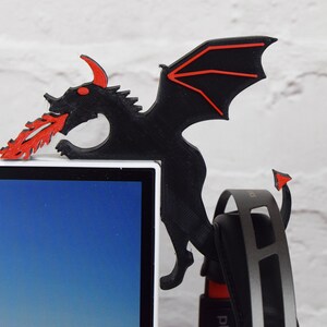 May include: A black dragon-shaped desk accessory with red accents, perched on the corner of a white computer monitor. The dragon has red eyes, horns, and a flame-like design emanating from its mouth. A pair of headphones hangs below.