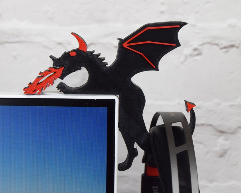 May include: A black dragon-shaped desk accessory with red accents, perched on the corner of a white computer monitor. The dragon has red eyes, horns, and a flame-like design emanating from its mouth. A pair of headphones hangs below.