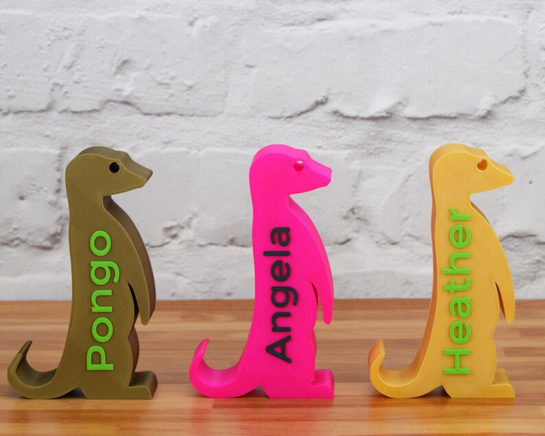 K&ouml;nnte beinhalten: Drei 3D-gedruckte Erdm&auml;nnchen-Figuren, jede in einer anderen Farbe: Braun, Pink und Gelb. Die Erdm&auml;nnchen stehen aufrecht mit ihren Schw&auml;nzen, die sich hinter ihnen einrollen. Jedes Erdm&auml;nnchen hat einen Namen auf der Seite gedruckt: Pongo, Angela und Heather.