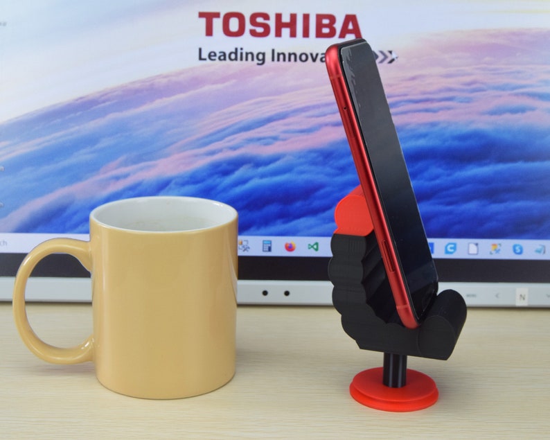 May include: A black 3D printed phone stand shaped like a hand holding a red smartphone. The stand is on a wooden surface next to a beige coffee mug.