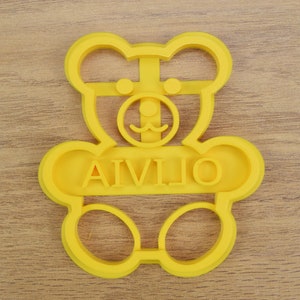Puede incluir: Cortador de galletas de pl&aacute;stico amarillo en forma de oso de peluche con el nombre "AIVILIO" escrito en el medio.