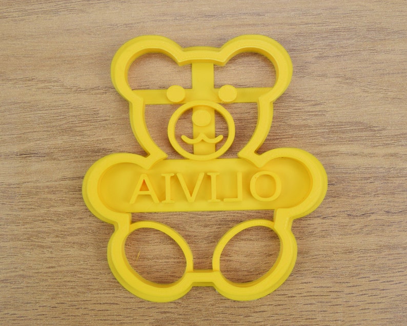Puede incluir: Cortador de galletas de pl&aacute;stico amarillo en forma de oso de peluche con el nombre "AIVILIO" escrito en el medio.