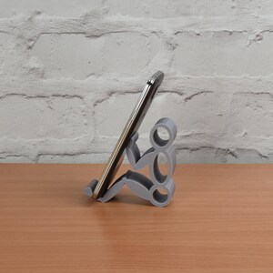 May include: A gray 3D printed phone stand shaped like a person holding a smartphone. The stand is on a wooden surface.