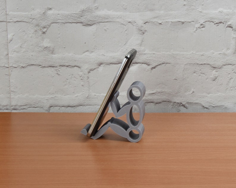 May include: A gray 3D printed phone stand shaped like a person holding a smartphone. The stand is on a wooden surface.