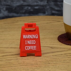 May include: A red warning sign with white text that reads "WARNING! I NEED COFFEE!" The sign is shaped like a traditional construction sign with two legs.