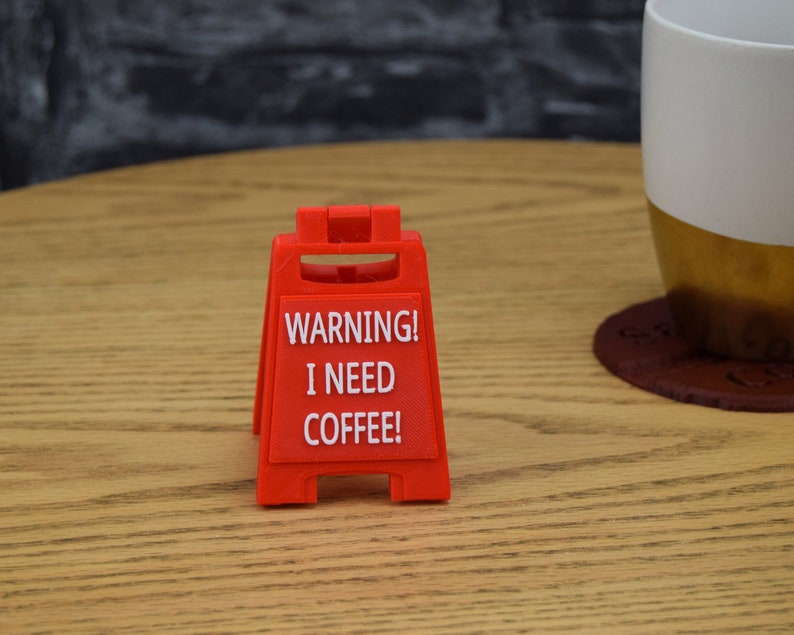 May include: A red warning sign with white text that reads "WARNING! I NEED COFFEE!" The sign is shaped like a traditional construction sign with two legs.