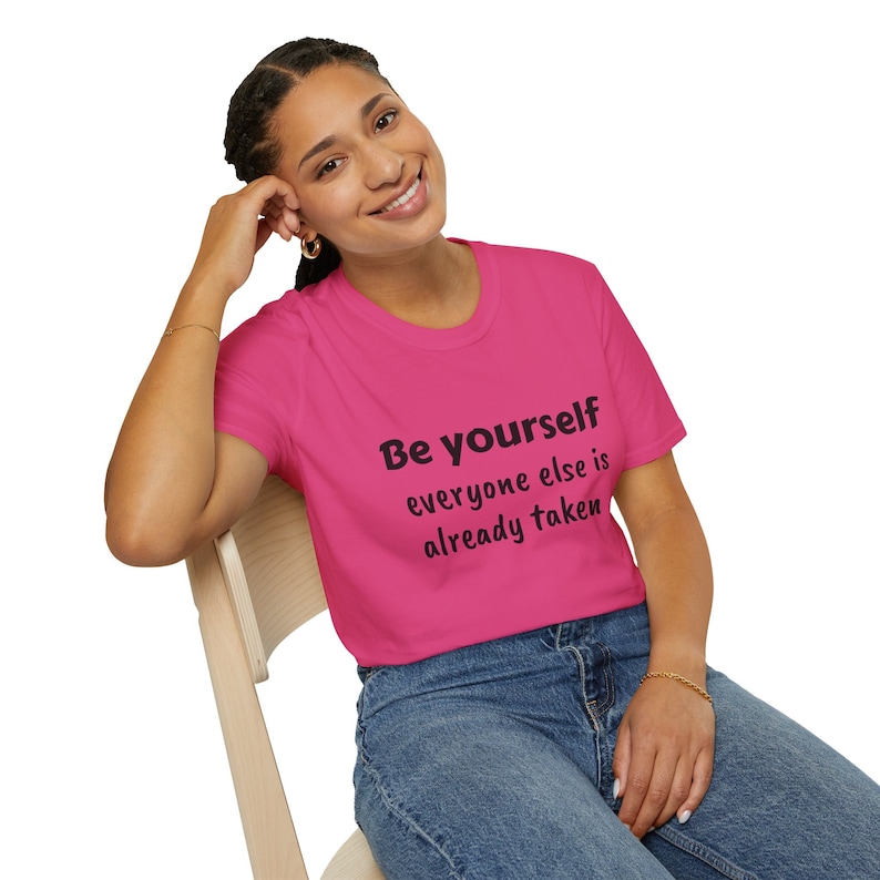 K&ouml;nnte beinhalten: Ein pinkfarbenes T-Shirt mit dem Text "Be yourself everyone else is already taken" in Schwarz.