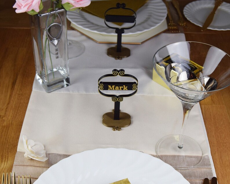 May include: A table setting with a white tablecloth, a glass vase with pink flowers, a martini glass, a white plate, and a black and gold place card holder with the name "Mark" on it.