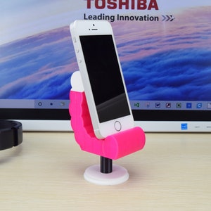 May include: A pink phone stand shaped like a chair with a white base. A smartphone is in the stand. The stand is on a wooden surface in front of a laptop screen.