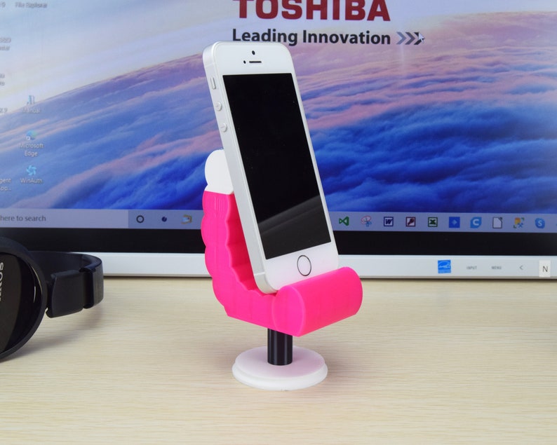 May include: A pink phone stand shaped like a chair with a white base. A smartphone is in the stand. The stand is on a wooden surface in front of a laptop screen.