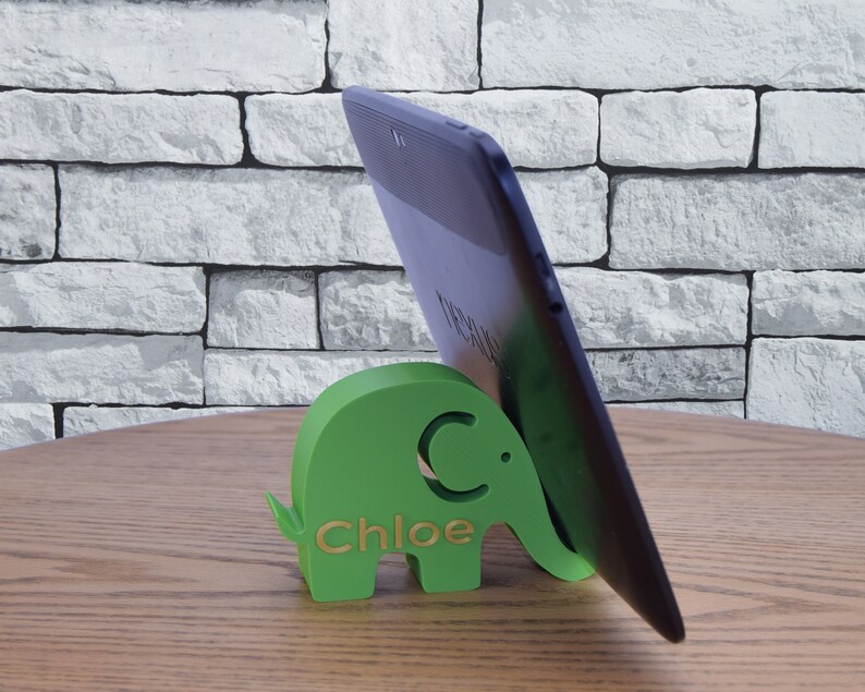 Pode incluir: Um suporte de telefone verde em forma de elefante com o nome "Chloe" gravado nele. Um tablet preto est&aacute; apoiado no suporte.