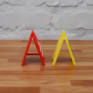May include: Two foldable road signs, one red and one yellow, are standing upright on a wooden surface. The signs are made of plastic and have a triangular shape.