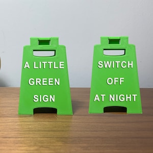 May include: Two green plastic signs with white text. The sign on the left says "A Little Green Sign". The sign on the right says "Switch Off At Night".