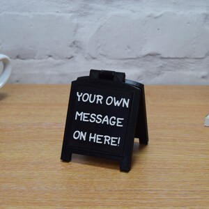 May include: A small, black A-frame chalkboard sign displaying the white text "YOUR OWN MESSAGE ON HERE!". The sign is on a wooden surface. A white coffee cup and a decorative house are visible in the background.