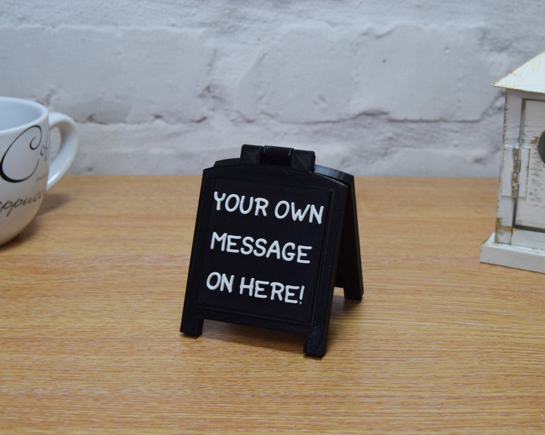May include: A small, black A-frame chalkboard sign displaying the white text "YOUR OWN MESSAGE ON HERE!". The sign is on a wooden surface. A white coffee cup and a decorative house are visible in the background.