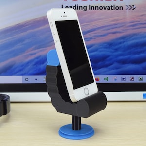 May include: A black and blue 3D printed phone stand shaped like a person sitting in a chair. The stand is holding a white iPhone.