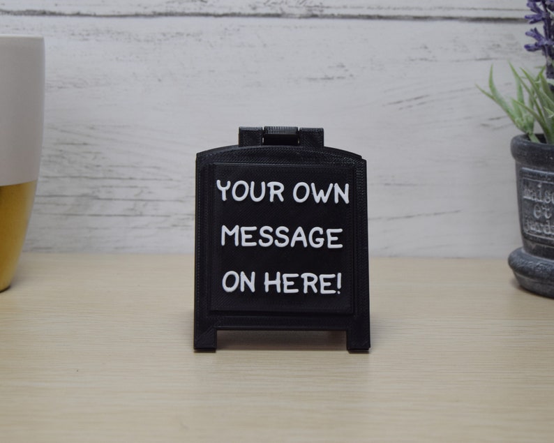 May include: A black chalkboard sign with the text "YOUR OWN MESSAGE ON HERE!" in white. The sign is in an A-frame style and sits on a light wooden surface. A decorative plant and a mug are in the background.