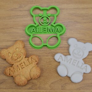 Puede incluir: Cortador de galletas de pl&aacute;stico verde en forma de osito de peluche con el nombre Amelia escrito en &eacute;l. Tambi&eacute;n se muestran dos galletas, una simple y otra glaseada.