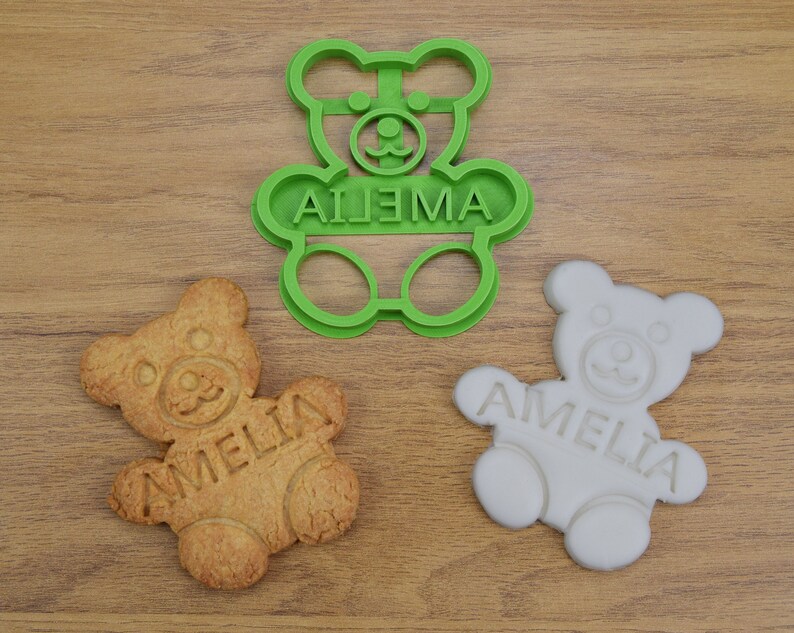 Puede incluir: Cortador de galletas de pl&aacute;stico verde en forma de osito de peluche con el nombre Amelia escrito en &eacute;l. Tambi&eacute;n se muestran dos galletas, una simple y otra glaseada.