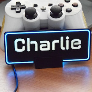 May include: A black sign with a blue neon border and the name "Charlie" in white text. The sign is sitting on a wooden surface.